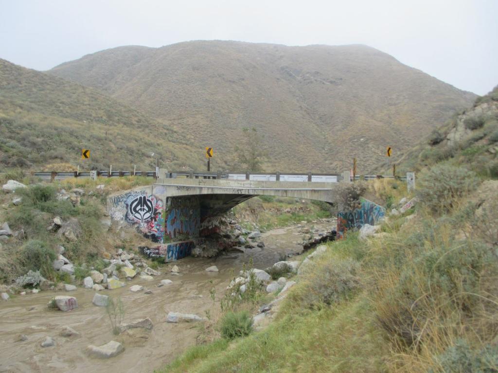 A small concrete bridge crosses a shallow, muddy stream in a dry, hilly landscape. The bridge and nearby rocks are covered in colorful graffiti. Yellow chevron road signs mark a curve on both sides of the bridge. The surrounding terrain has sparse shrubs, dry grass, and rocky slopes under an overcast sky.